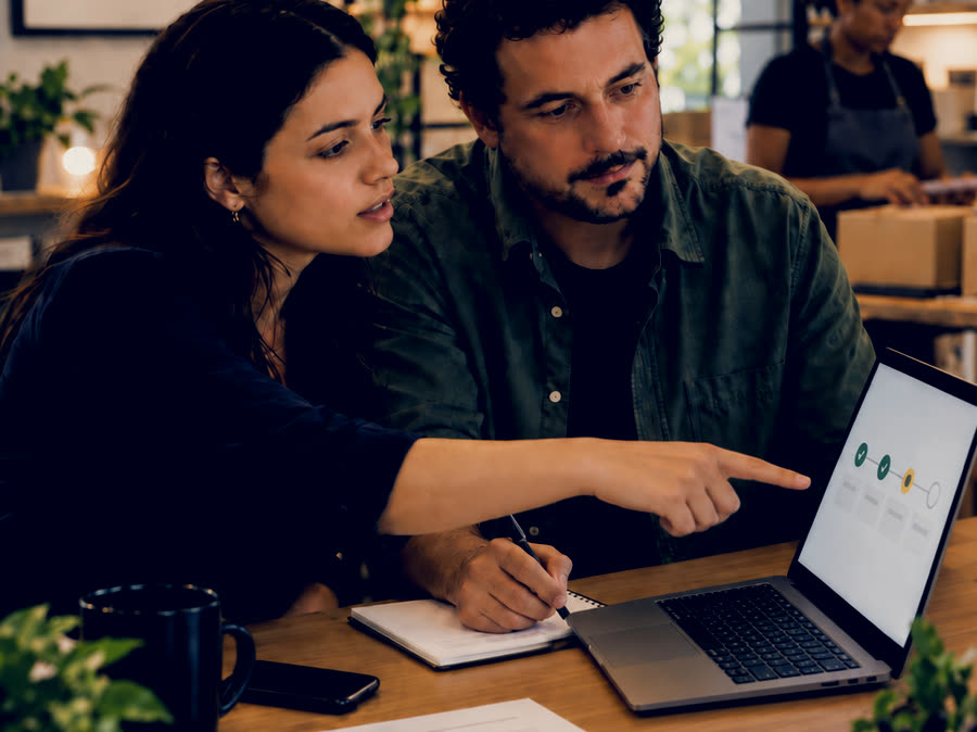 Duas pessoas acompanham o fluxo rodando na tela de um notebook, com etapas marcadas em verde e uma em amarelo pedindo atenção; uma delas aponta para a tela enquanto a outra anota os ajustes.