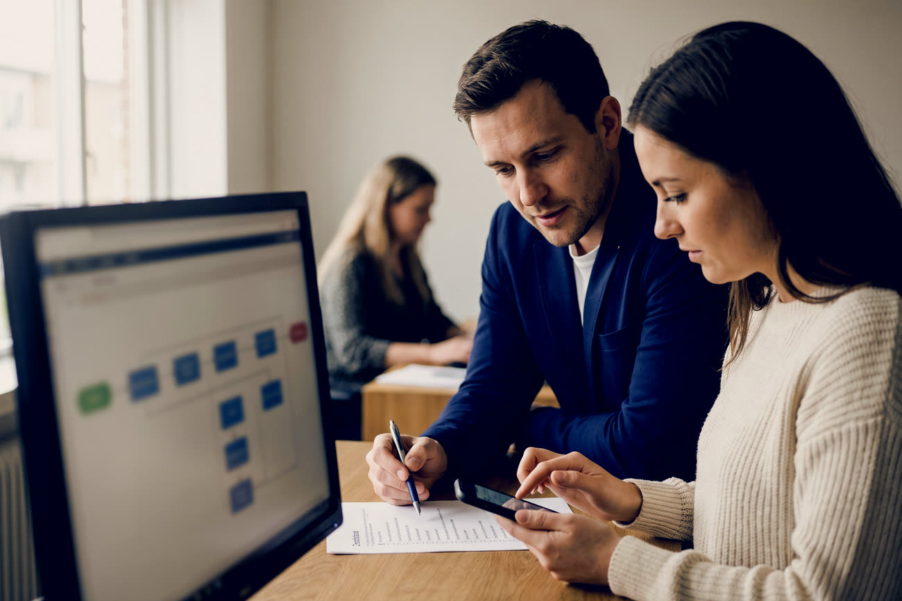 Duas pessoas lado a lado revisando um fluxo de processo exibido no monitor, enquanto uma anota itens em uma lista de verificação e a outra confere algo no celular.
